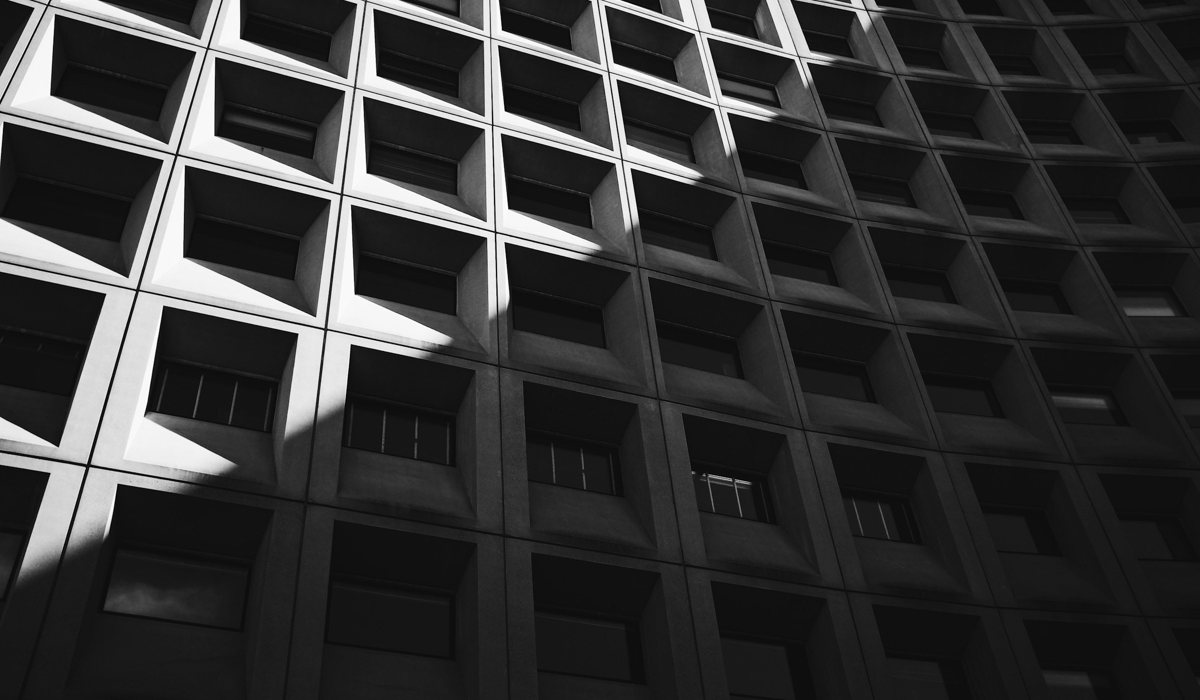Sort-hvidt foto af en betonfacade set nedefra, hvor ens vinduesfelter går igen række efter række, og hvert felt står forskelligt i lys, skygge og persienner.